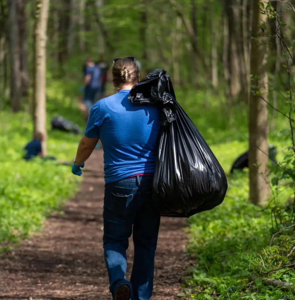 A person wearing a blue shirt and gloves carries a large black garbage bag over their shoulder while walking on a forest trail. Other people are visible in the background, participating in the cleanup effort. Lush greenery surrounds the path.