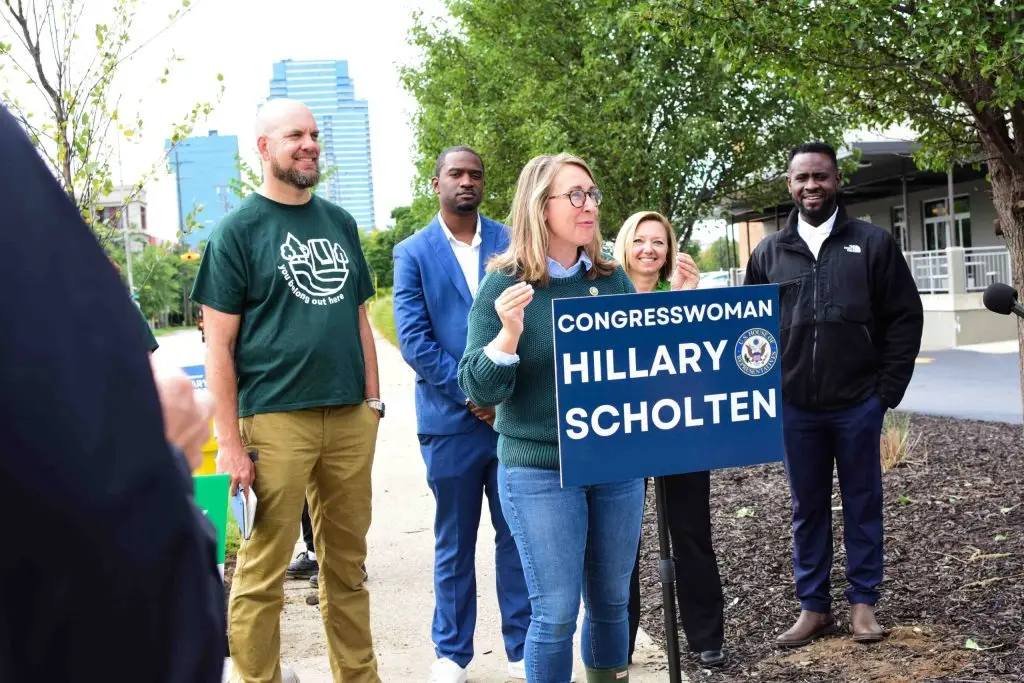 A group of five people stands outdoors, with one person holding a sign that reads Congresswoman Hillary Scholten. They are surrounded by greenery and city buildings are visible in the background.