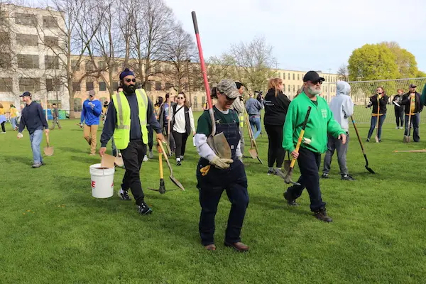 A group of people wearing casual and work clothes walk on a grassy field carrying shovels and a bucket. Trees and buildings are in the background. The event appears to be related to planting or gardening.