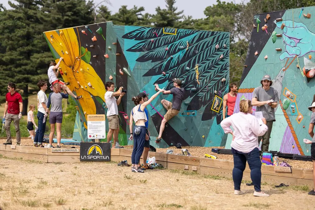 People climbing colorful outdoor bouldering walls with a crowd watching. The walls feature vibrant designs, including a yellow figure with flowers and a black feather. Climbers are focused, and the scene is lively with observers in casual attire.