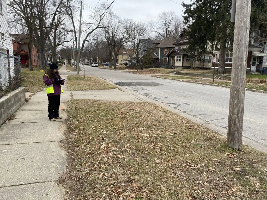 A person in a purple jacket and yellow vest stands on a sidewalk, using a device. The street is quiet with bare trees and residential houses. Its a cloudy day, and the grass appears brown and sparse.