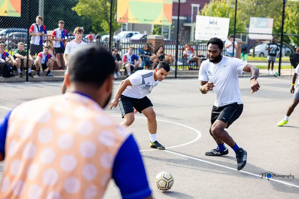 A group of people play soccer on an outdoor court. Two players in white shirts focus on the ball, with one approaching it. Spectators watch from the background behind a fence. Its a sunny day, and the scene is lively and energetic.
