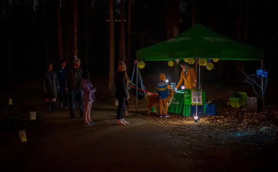 People are gathered under a green tent lit by hanging lanterns in a dark forest. A few individuals and a dog are lined up near a table with a person assisting them. Paper bags with lights line the path.