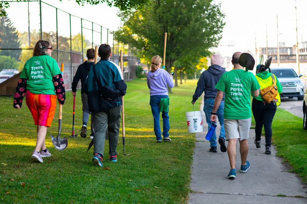 A group of people walking on a sidewalk next to a grassy area. Some are carrying tools like shovels and buckets. Two people wear green shirts with the words Trees, Parks, Trails. The scene suggests a community or volunteer event.