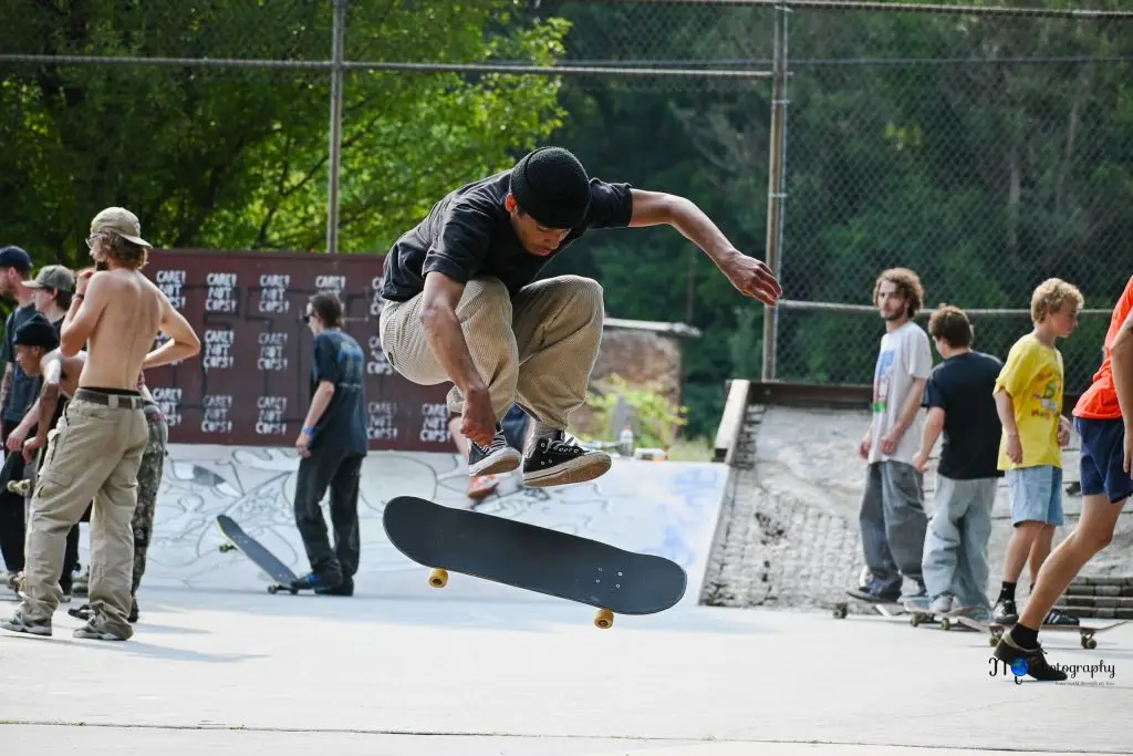 A skateboarder performs a trick mid-air at a skatepark, surrounded by several people watching. The background features graffiti and a chain-link fence.
