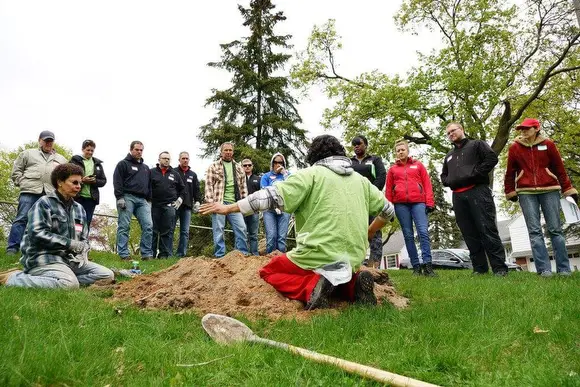A group of people standing in a semi-circle around a person kneeling with arms outstretched on a grassy area. A shovel is placed on the grass in front. Trees are visible in the background.