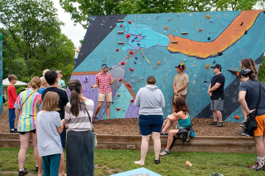 A group of people gathers outdoors to watch a man demonstrate climbing on a colorful mural-covered bouldering wall. The mural features large painted arms and hands. Trees and grass are visible in the background.